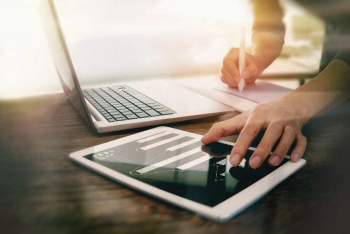 Accountant reviewing a transaction on an iPad, writing T-accounts using the DEALER rule, with a laptop ready for digital entry